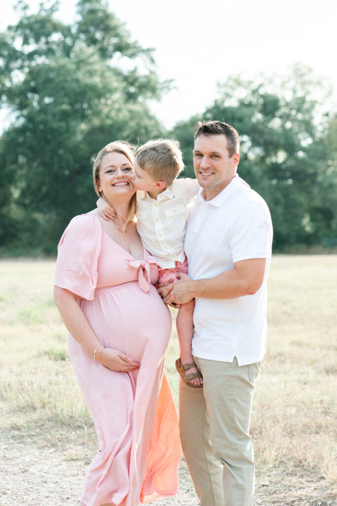 A family of three stands on a sandy path. The father holds his son while the son wraps his arms around his mother's neck and kisses her on the cheek. The pregnant mother, in a long pink maternity dress, holds her belly and smiles in a tender moment beautifully captured by an Austin maternity photographer.