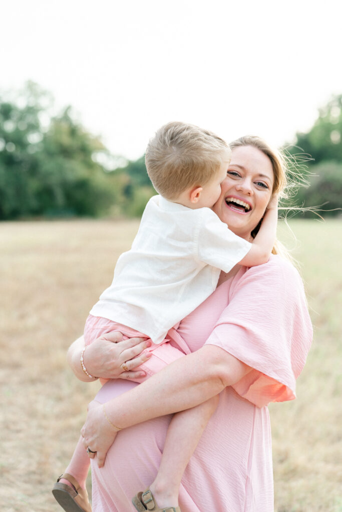 A pregnant mother in a long pink maternity dress stands on a sandy path while holding her son dressed in a white shirt and pink shorts. The son wraps his arms around his mother's neck to hug her while the mother smiles in a tender moment captured by an Austin maternity photographer.