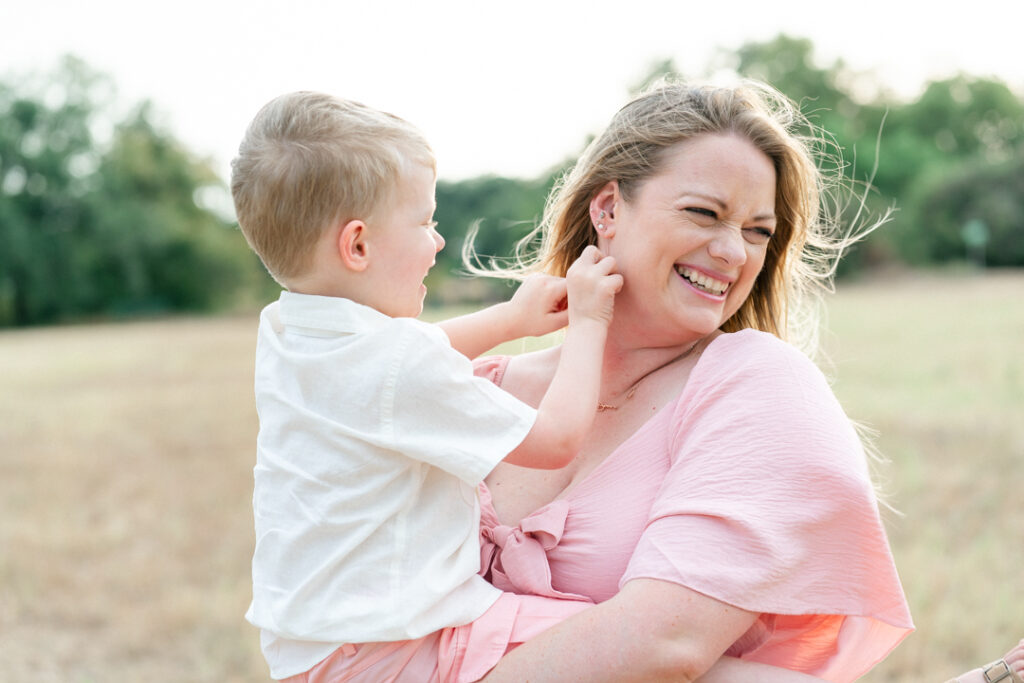 A pregnant mother in a long pink maternity dress stands on a sandy path while holding her son dressed in a white shirt and pink shorts. The son tickles his mother while the mother looks away and laughs in a tender moment captured by an Austin maternity photographer.