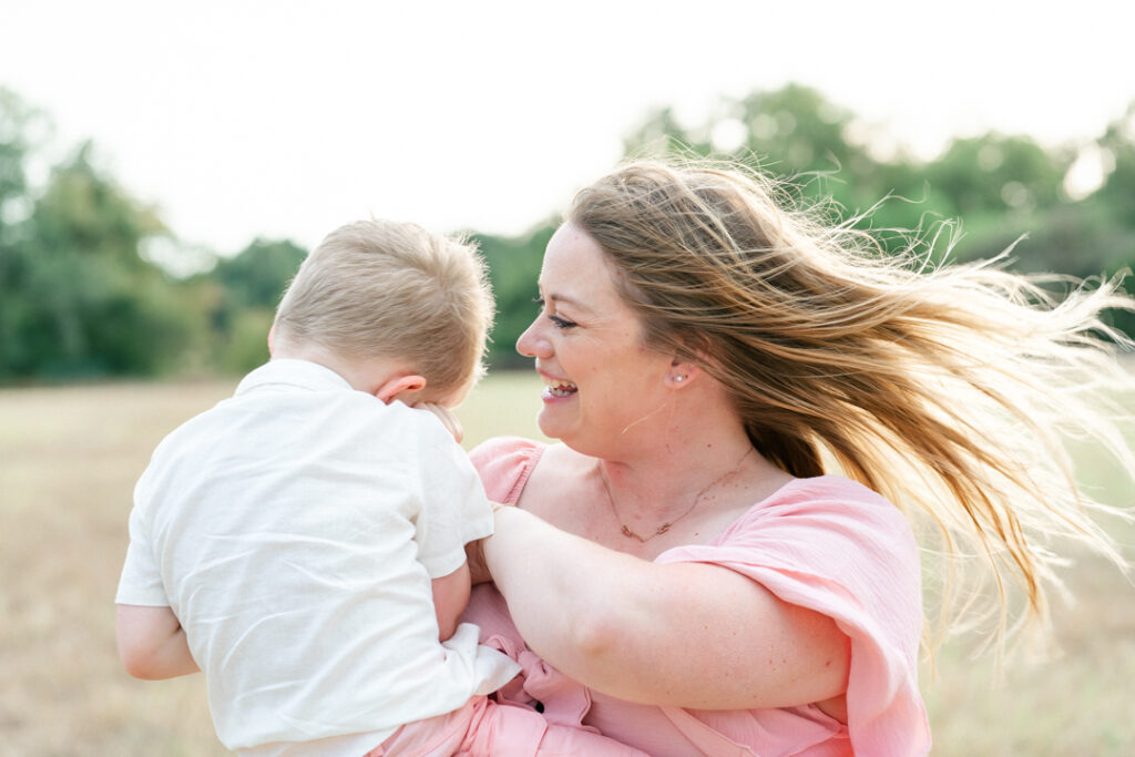 A mother in a pink dress holds her son dressed in a white shirt and pink shorts. The mother tickles her son and laughs in a tender moment captured by an Austin maternity photographer.