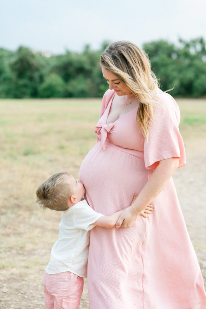 A pregnant mother in a long pink maternity dress stands on a sandy path. Her son stands facing her and kisses her pregnant belly while the mother looks down at him with a smile in a tender moment captured by an Austin maternity photographer.