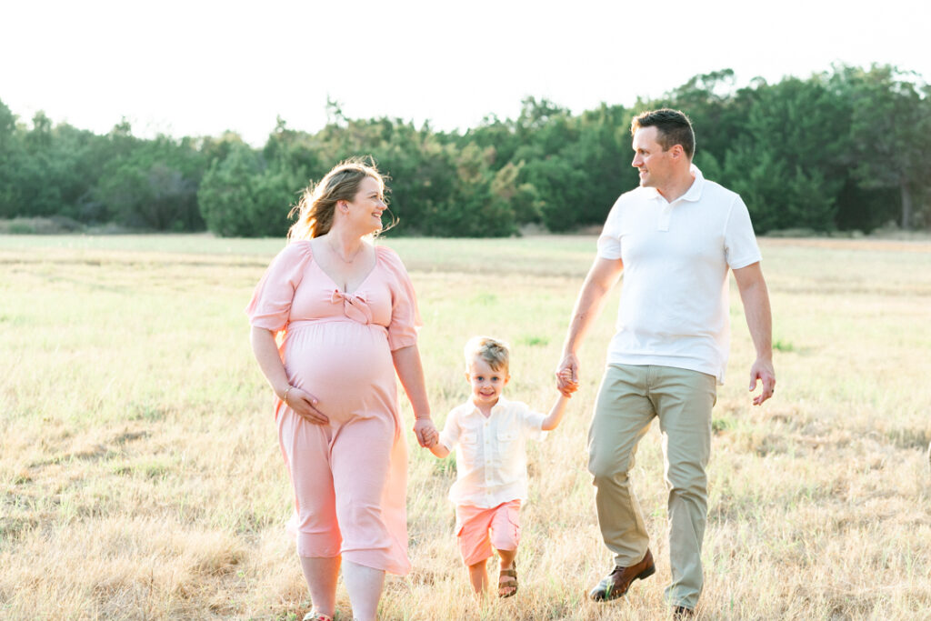 A family of three walks in a grassy field with green trees in the background. The mother, in a pink maternity dress, holds her belly while looking at her husband. The son, in the middle, holds his mother and father's hand and smiles, beautifully captured by an Austin Maternity photographer.