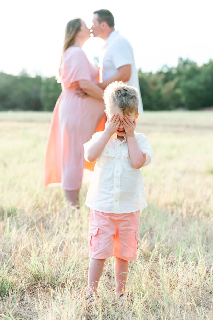 A young boy stands in a grassy field and covers his eyes with his hands while his mother and father stand in the background. The mother, in a long pink maternity dress, and the husband kiss while the husband holds his wife's pregnant belly, in a sweet and moment captured by an Austin maternity photographer.