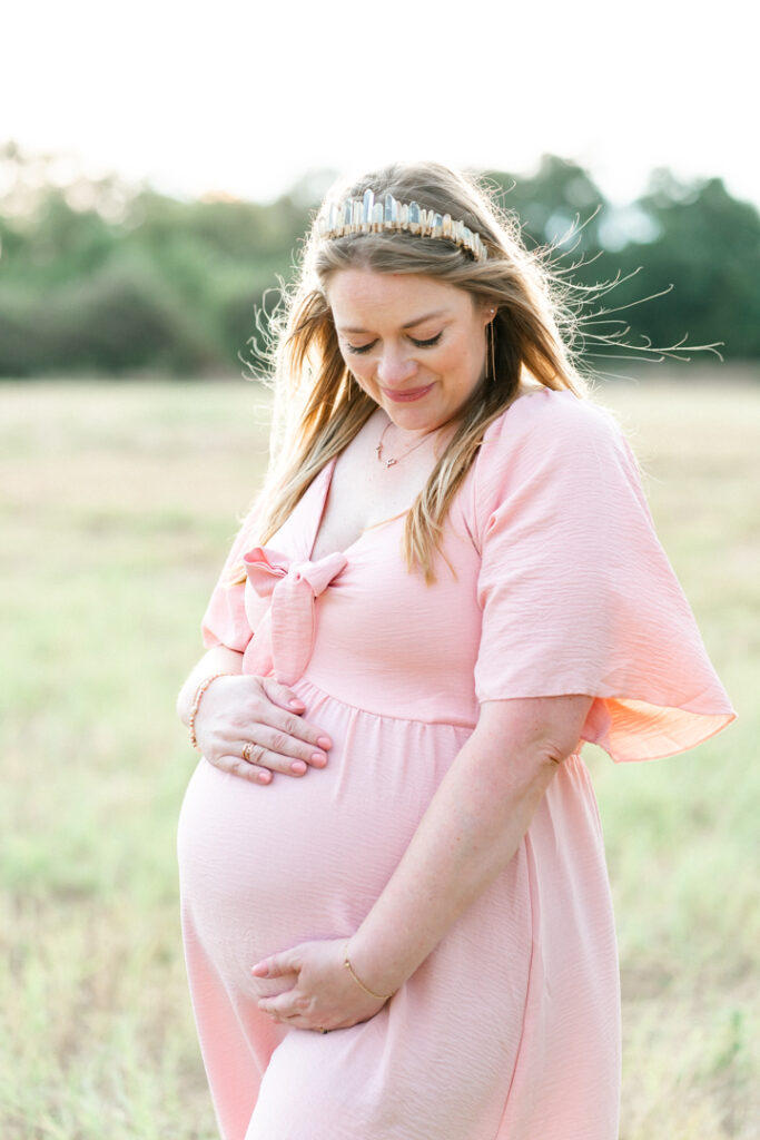 A pregnant mother in a soft pink dress stands in a park. She looks down at her belly in this tender photo captured by an Austin Maternity Photographer