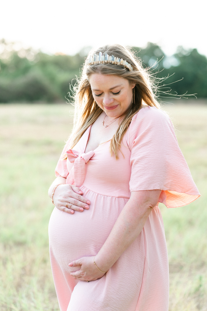 A pregnant woman dressed in a pink maternity dress stands is a grassy field. She wears a crown in her blond hair and looks down at her belly while smiling. She holds her belly with both hands in a tender moment captured by an Austin maternity photographer.