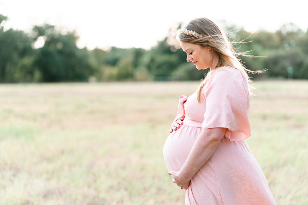 A pregnant woman dressed in a pink maternity dress stands is a grassy field. She wears a crown in her blond hair and looks down at her belly while smiling. She holds her belly with both hands in a tender moment captured by an Austin maternity photographer.