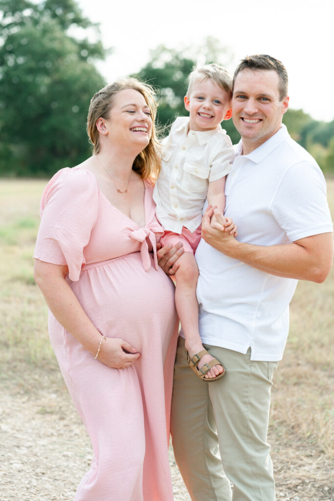 A family of three stands on a sandy path. The father, dressed in a white shirt, holds his son in his arms. The son holds his father's hand. The pregnant mother, in a long pink maternity dress, holds her belly and smiles with her eyes closed in a tender moment beautifully captured by an Austin maternity photographer.