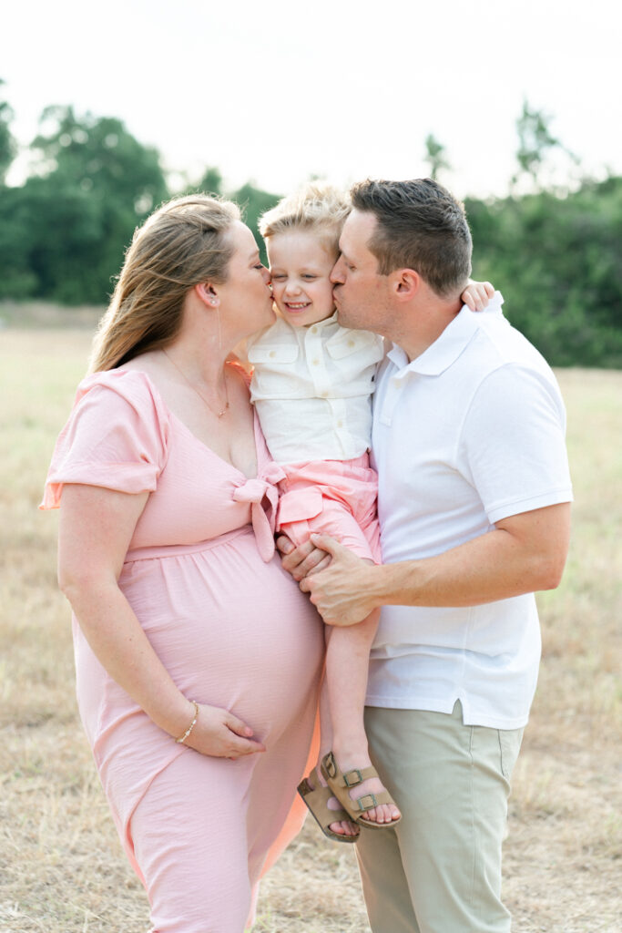 A family of three stands on a sandy path. The father and mother hold their son in between them while kissing him on the cheeks. The pregnant mother, in a long pink maternity dress holds her belly, captured beautifully by an Austin maternity photographer.