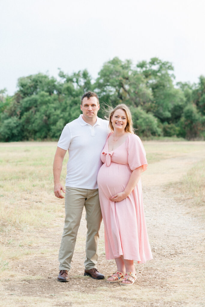 A wife and husband stand on a sandy path. The husband, dressed in a white shirt and kaki pants, wraps his arm behind his wife. The pregnant wife, in a long pink maternity dress, smiles while holding her belly, captured beautifully by an Austin maternity photographer.