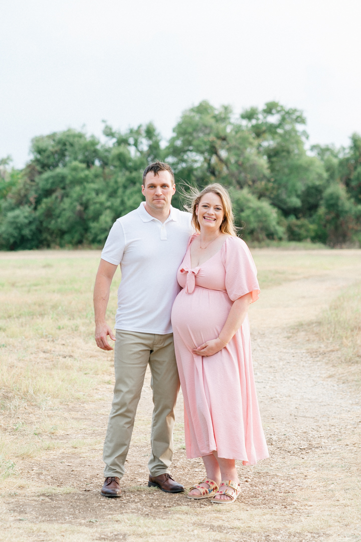 A wife and husband stand on a sandy path. The husband, dressed in a white shirt and kaki pants, wraps his arm behind his wife. The pregnant wife, in a long pink dress, smiles while holding her belly, captured beautifully by a Cedar Park photographer.