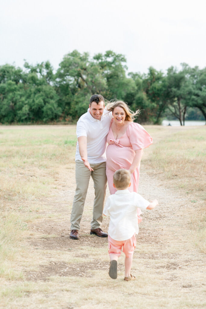 A father and a pregnant mother are standing on a sandy path while looking and smiling at their son running towards them, captured beautifully by an Austin maternity photographer.