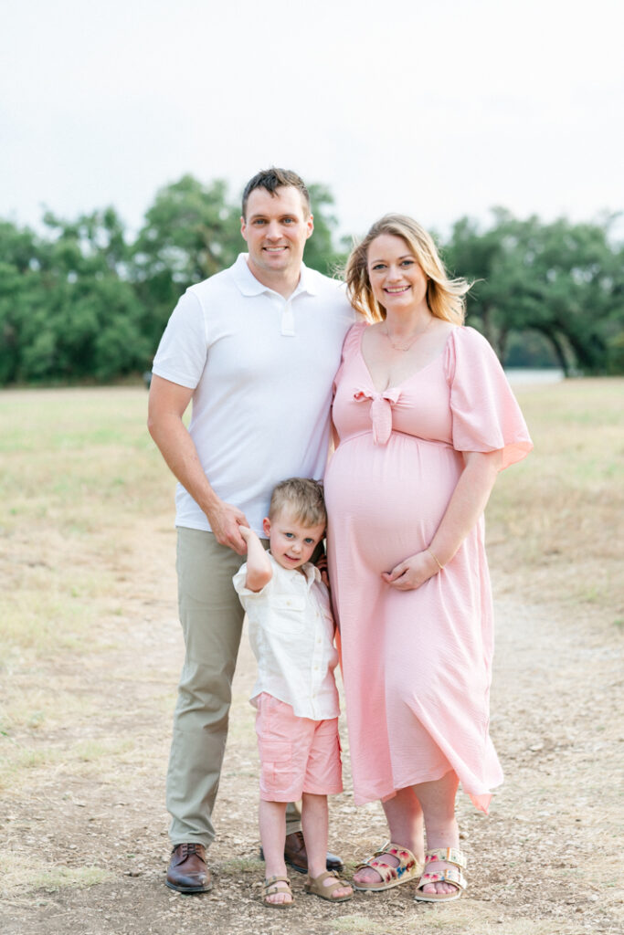 A family of three stands on a sandy path. The father wraps his arm around his pregnant wife's waist, while the son stands in between his parents and holds his father's hand, in a joyful moment captured by an Austin maternity photographer.