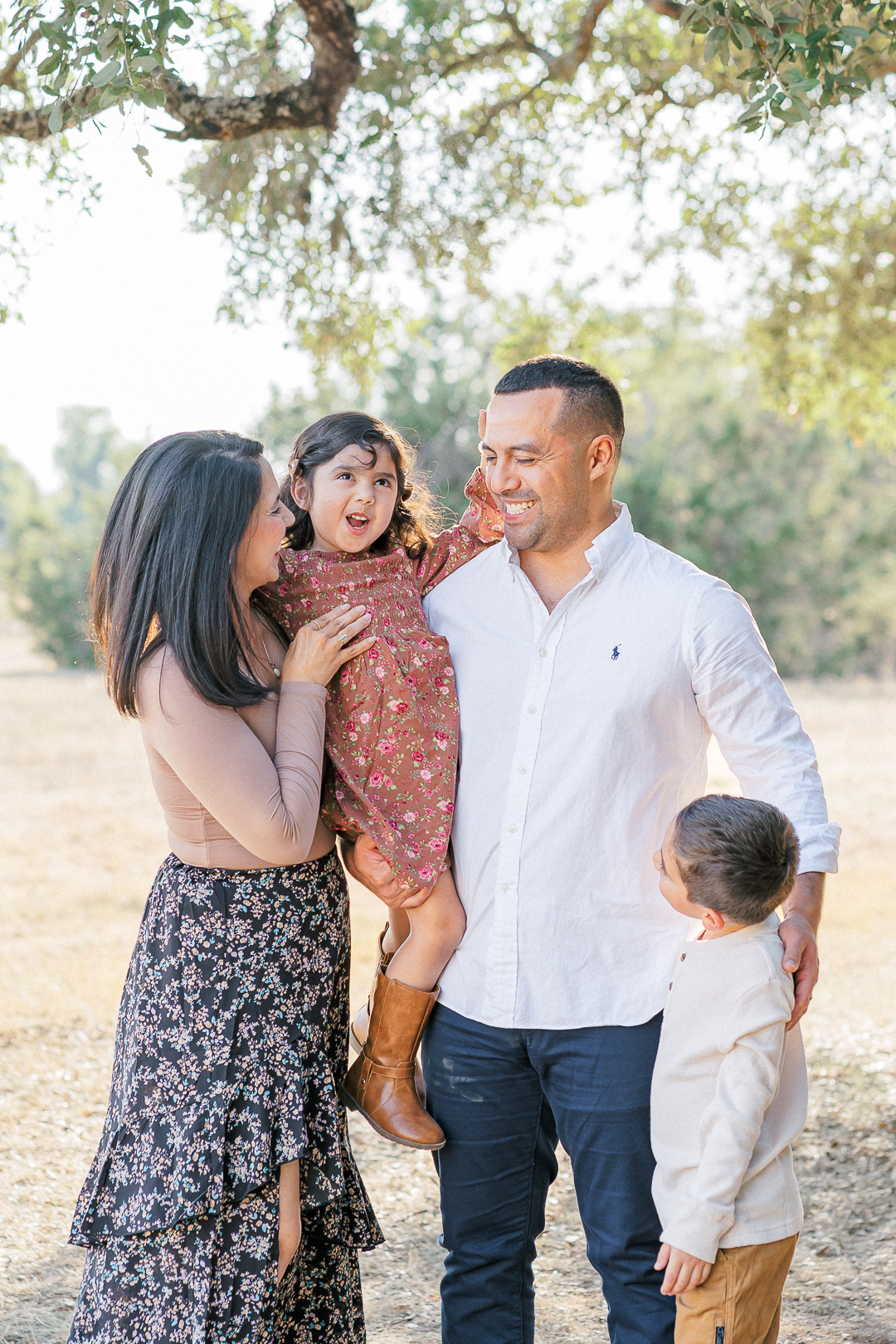 A family of four stands outdoors under a tree, captured perfectly by an Austin family photographer. The mother holds a young girl in a floral dress, while the father, in a white shirt, stands beside them. A young boy in a light sweater and dark pants looks up at him. The peaceful setting embodies Texas charm.