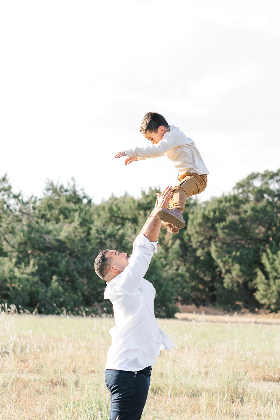 A man in a white shirt joyfully lifts a young boy in light clothing into the air, captured beautifully by an Austin family photographer. They are outdoors in a grassy field with tall trees silhouetted against the clear sky.