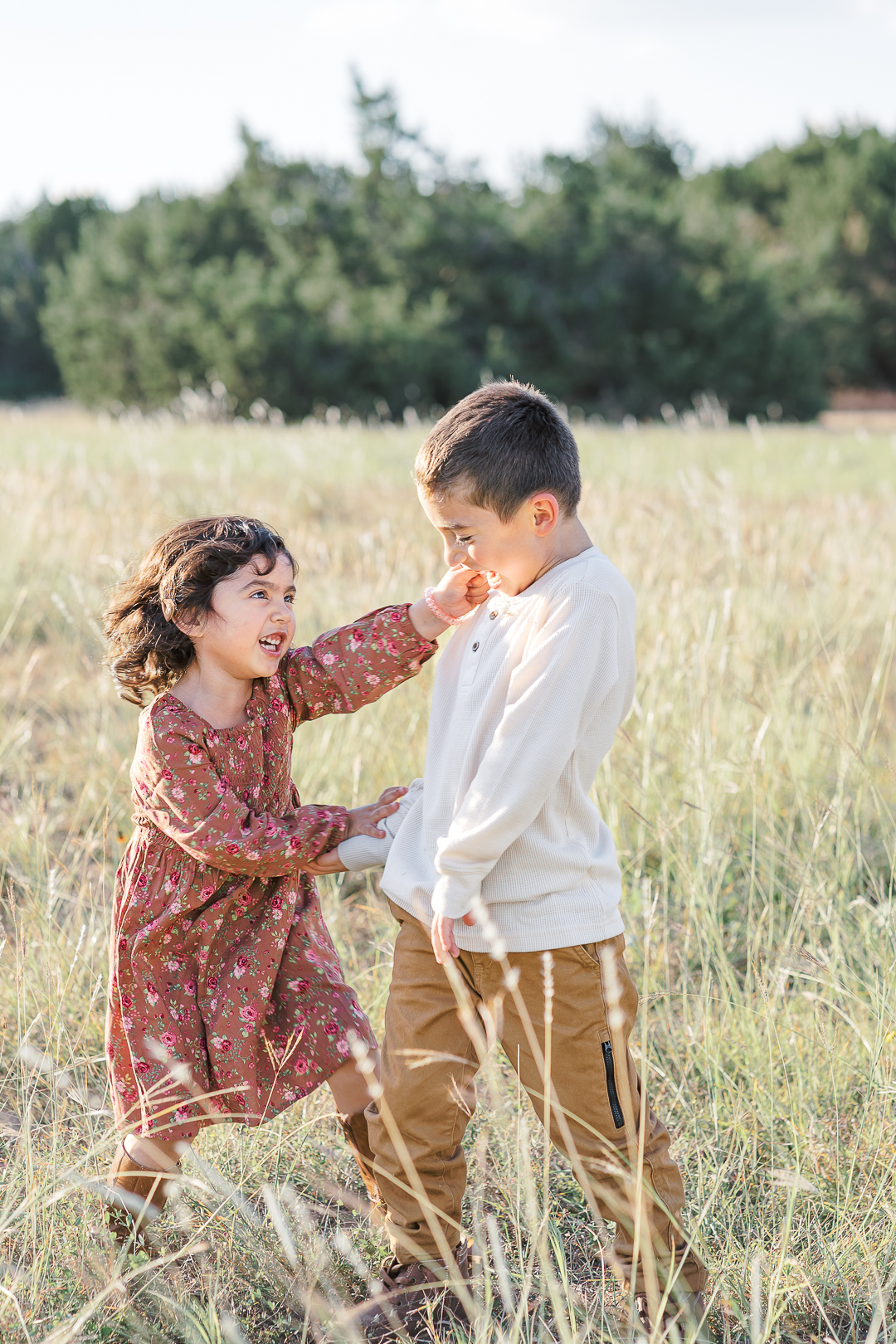 A girl in a brown floral dress is playing with a boy in a white shirt in a grassy field. The girl and the boy are facing each other and laughing, in an authentic moment captured beautifully by an Austin family photographer.