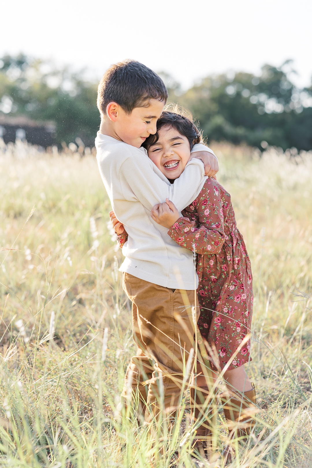 A young boy in a white short is hugging a young girl in a floral dress while standing in a grassy field. The boy and the girl have their eyes closed and the girl is smiling, a timeless moment beautifully captured by an Austin family photographer.