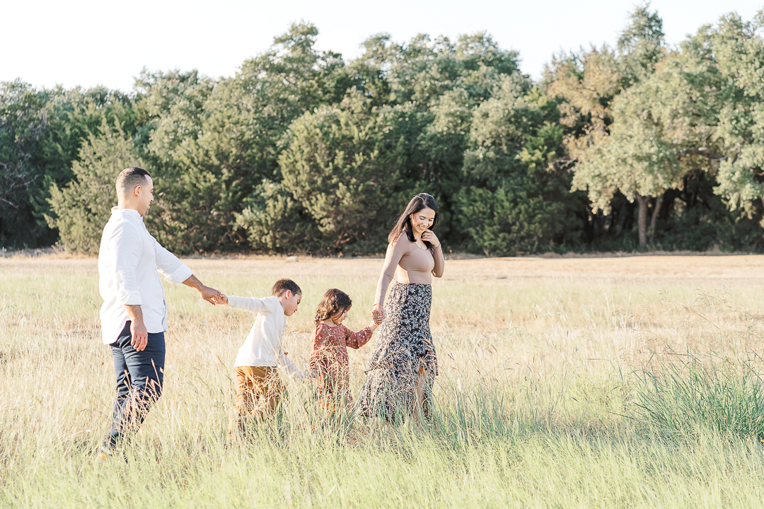 A family of four walks in a grassy field with the mother leading the way. Her daughter, son, and the father are following behind her. The mother looks down at her daughter while smiling, beautifully captured by an Austin family photographer.