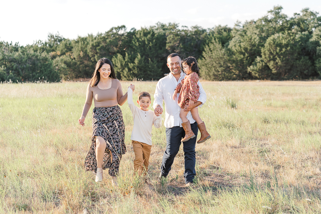 A family of four walks in a grassy field. The son is in the middle and holds his mother and father's hand. The mother, son, and father smile while walking while the daughter, in her father's arms, looks down at her brother in a timeless moment beautifully captured by an Austin family photographer.