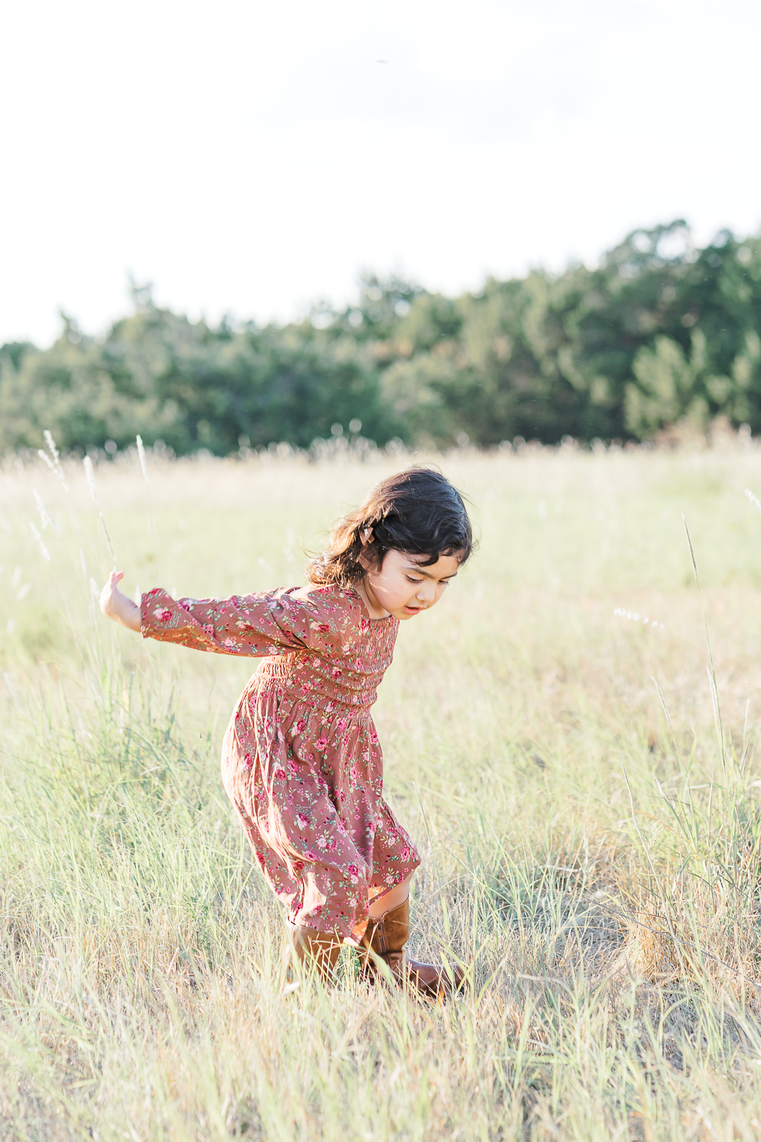 A young girl in a floral dress is jumping in a grassy field with green trees in the background, beautifully captured by an Austin family photographer.