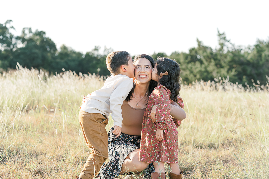 A mother kneels in a grassy field while hugging her son and daughter on each side of her. The son and daughter kiss their mother on the cheeks while the mother looks at an Austin family photographer's camera and smiles. 