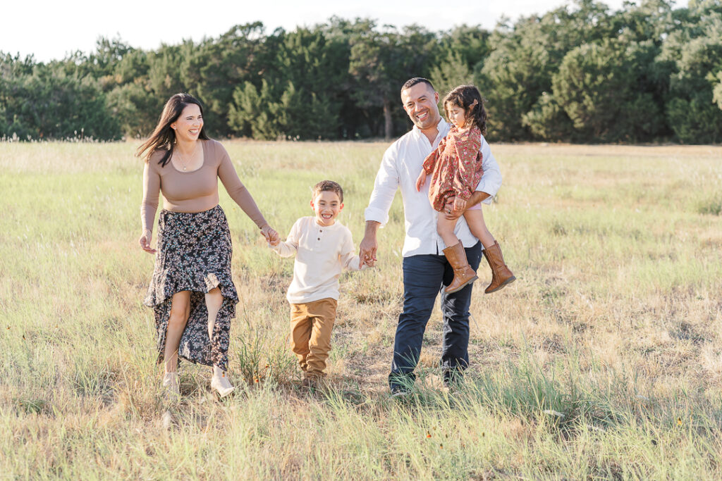 A family of four walks through a grassy field in Austin,TX. The mother holds the son's hand, and the father carries the daughter. Trees are in the background.