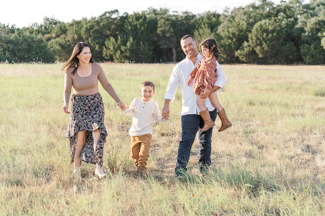 A family of four walks through a grassy field. The mother holds the son's hand, and the father carries the daughter. Trees are in the background.