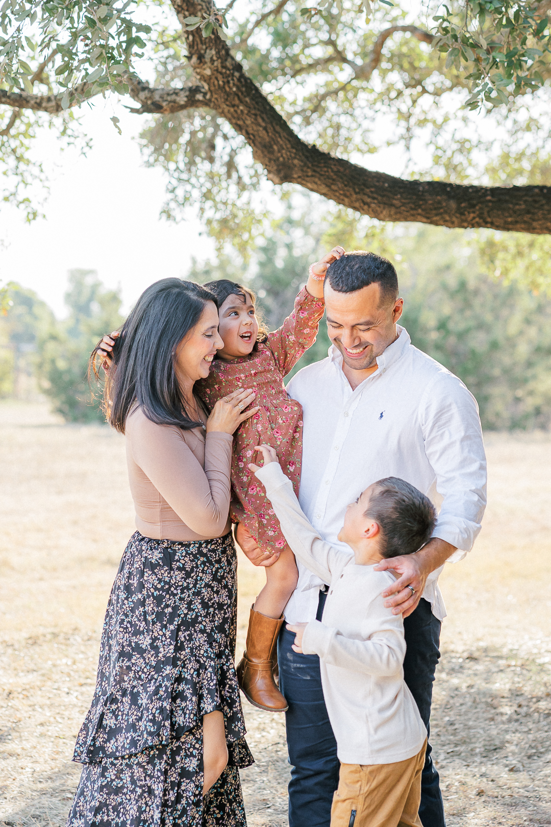 A family of four stands under a tree in a sunny park, captured beautifully by an Austin family photographer. The family is looking at each other laughing. The father holds the daughter while she tickles his head and touches her mother's hair. The brother tickles his sister while his parents look down at him laughing.