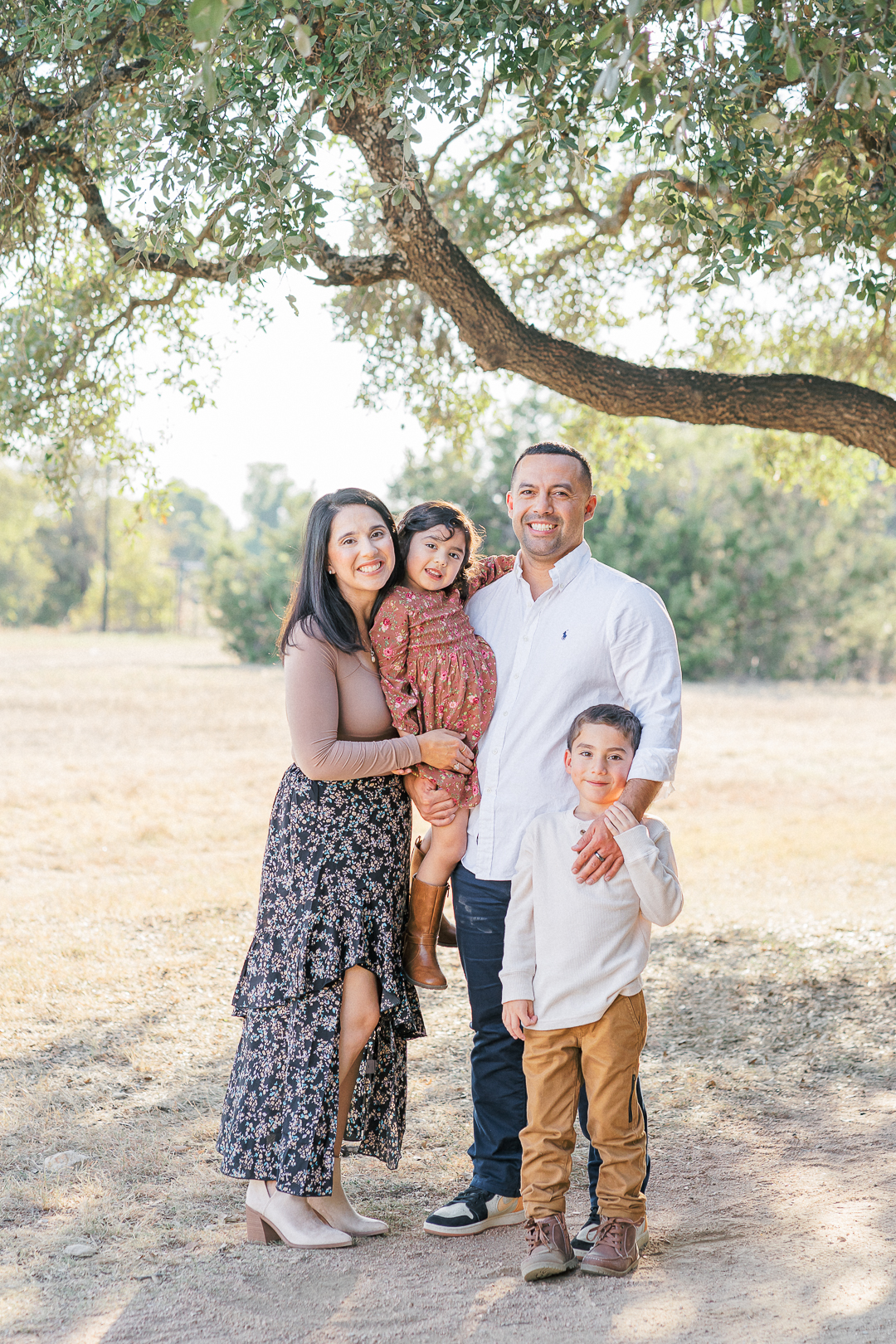 A family of four stands under a tree in a sunny park, captured beautifully by an Austin family photographer. The parents smile at the camera, holding their young daughter, while their son stands beside them. The background features lush grass and trees.