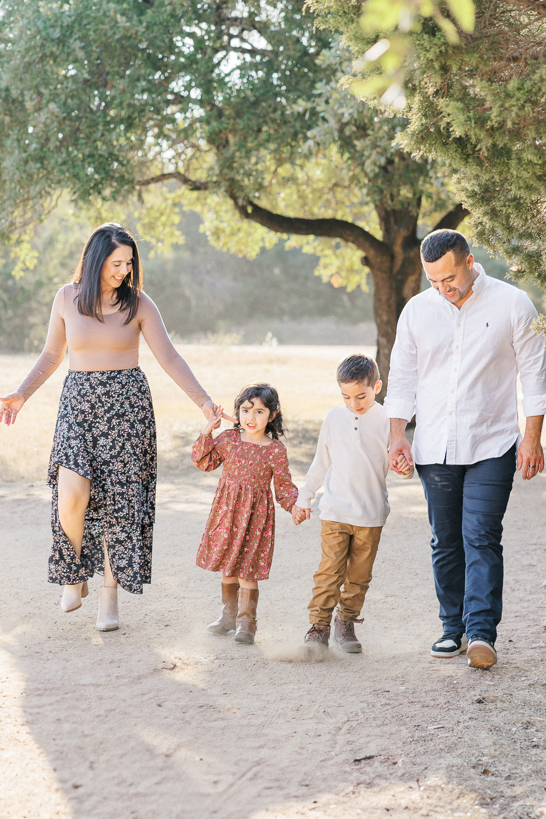 A family of four is walking down a sandy path in a park, captured beautifully by an Austin family photographer. The mother, father, and son look down at their feet smiling while the daughter looks ahead. There is beautiful light peaking through a tree in the background.