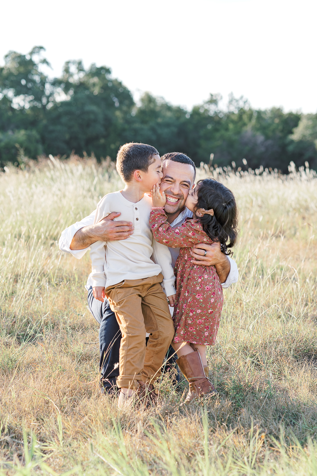 A man kneels in a grassy field, joyfully hugging a young boy and girl. The boy, in a white shirt and brown pants, kisses him on the cheek while the girl, wearing her floral dress and boots, stands beside them. The authentic moment is captured beautifully by an Austin family photographer amidst the backdrop of trees.