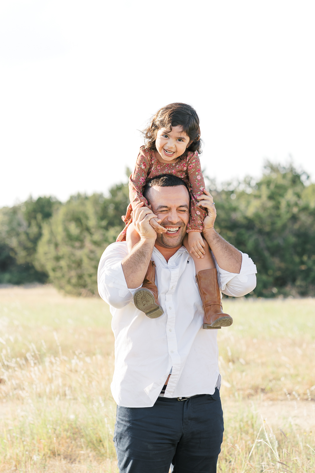 An Austin family photographer captures a timeless and genuine moment: a smiling man carries a joyful child on his shoulders in a grassy field. The child laughs while holding onto the man's head, as he playfully grips the child's legs, with trees under a bright sky in the background.