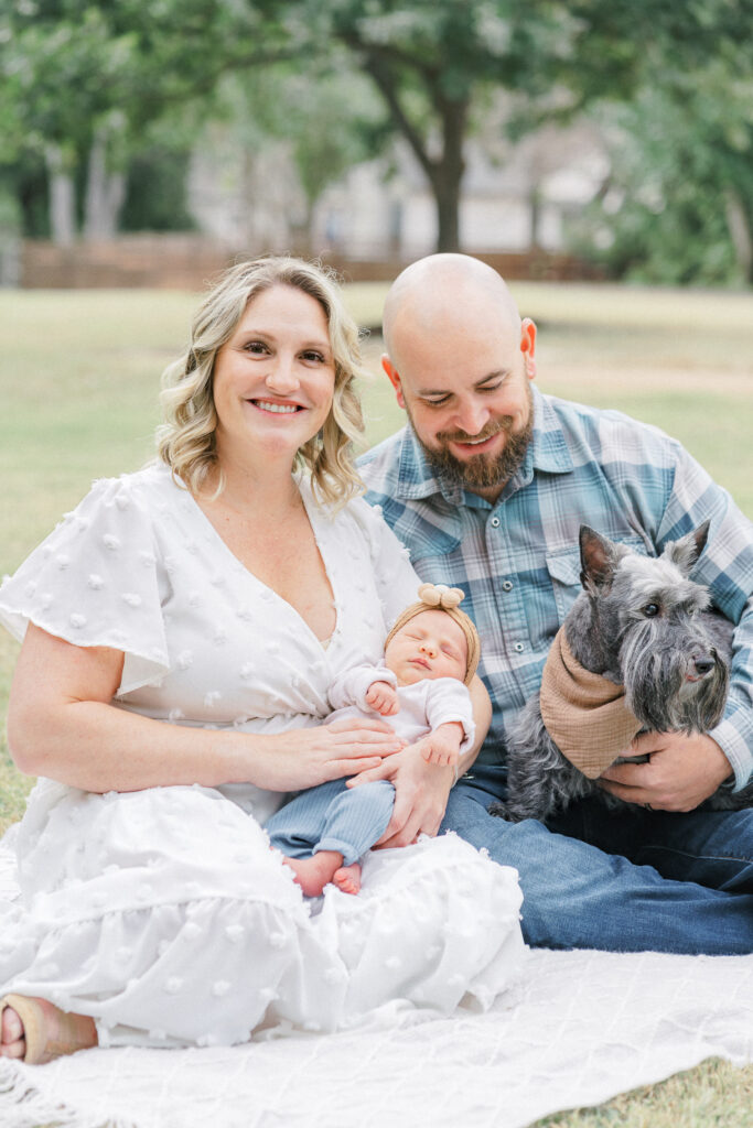 A mother in a long white dress sits on a white blanket in a park while holding her newborn baby. Her husband sits next to her and holds their grey dog. The father looks down at their newborn baby and smiles. The mother smiles at the Austin Newborn Photographer's camera.