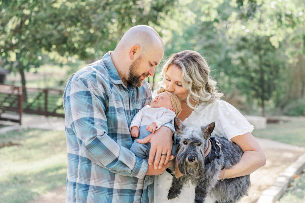 A mother kisses her newborn baby girl on her head while holding their grey dog. The father holds the newborn baby and looks down at her smiling in this tender outdoor newborn shot captured by an Austin Newborn Photographer.