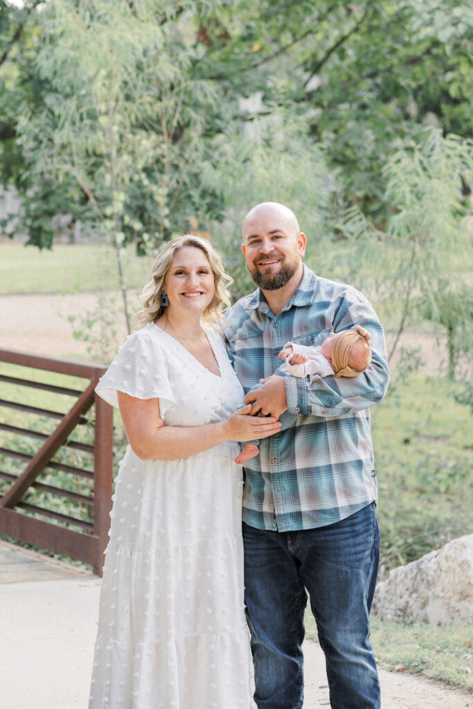 A father holds his newborn baby and smiles at the camera. His wife stands next to him with her hand on their newborn baby and smiles at the camera during an outdoor newborn photo session captured by an Austin Newborn Photographer.