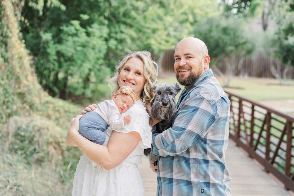 A mother holds her newborn baby girl with her head resting on her shoulder. Her husband stands behind her holding their grey dog, at their outdoor newborn photo session captured by an Austin Newborn Photographer