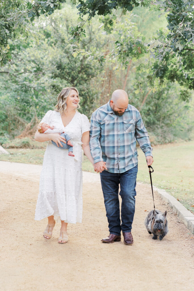 A mother and father walk on a sandy path in a park while holding hands. The mother holds their newborn baby while the father holds their dog on a leash. They smile in this genuine moment captured by an Austin Newborn Photographer.