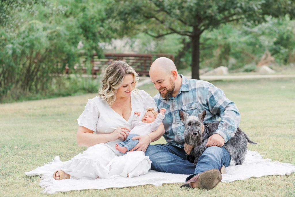 A family sits on a blanket in the park, captured by an Austin newborn photographer. The light and airy scene features a man in a plaid shirt holding their baby alongside a woman in a white dress. A small dog sits by them, with trees and grass providing a serene backdrop for this outdoor newborn photo session.