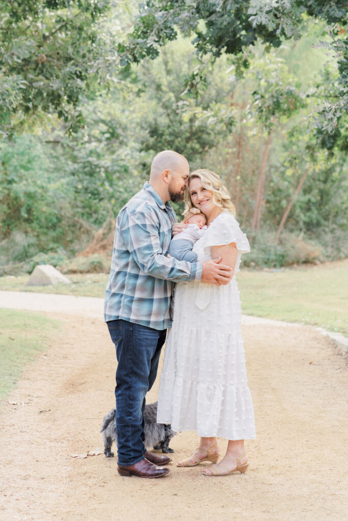 A mother and father stand on a sandy path in a park. The mother holds their newborn baby. The father rubs his nose on the temple of the mother's head. The mother, in a long white dress, smiles at the camera of an Austin Newborn Photographer. Hidden behind them is their grey haired dog. 