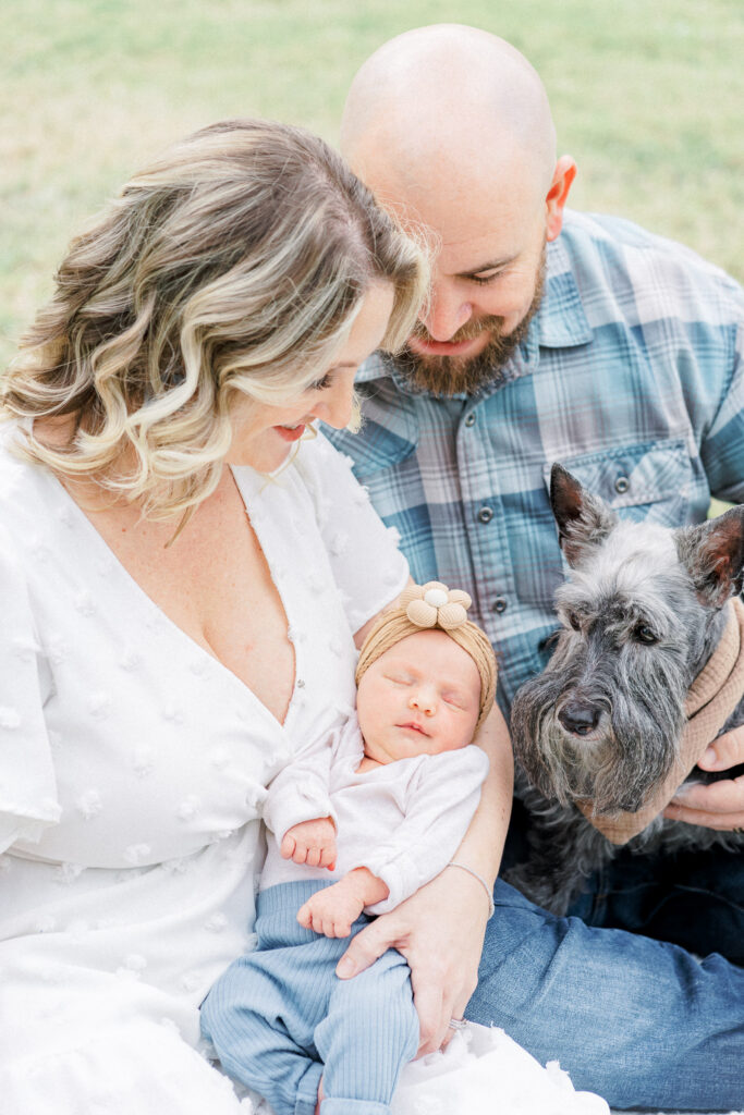 A mother and father sit in a park while the mother holds their newborn baby girl. The parents look down at their newborn baby while smiling. The father holds they grey haired dog. The baby is peacefully sleeping in her mother's arms in this beautiful shot captured by an Austin newborn photographer.