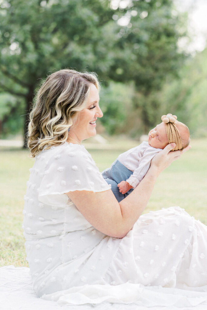 A mother sits on a white blanket and holds her newborn baby. She looks down at her baby smiling, in a tender moment captured by an Austin Newborn Photographer.