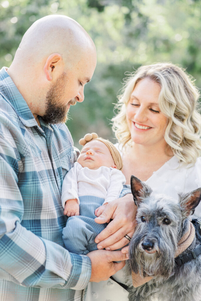 A mother and father look down smiling at their newborn baby. The father holds the baby while the mother holds their grey dog, in a joyful moment captured by an Austin Newborn Photographer.