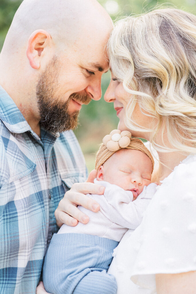 A mother and father face each other and lean their foreheads against one another while the mother holds their newborn baby in between them in a sweet outdoor newborn photo session shot by an Austin Newborn Photographer.