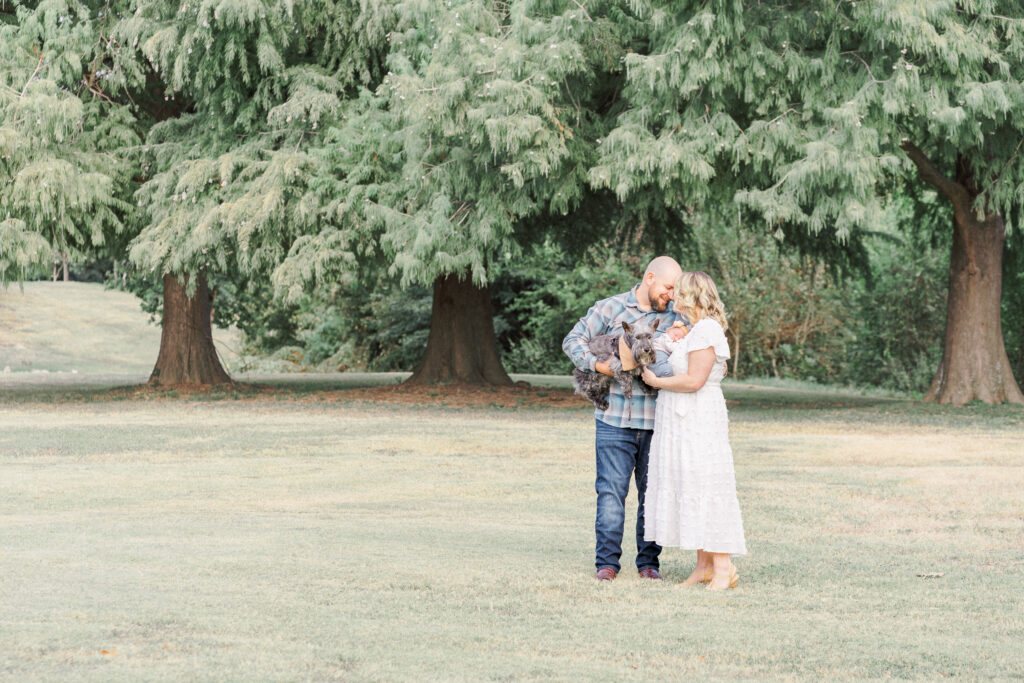 A mother and father have their forehead together while the mother holds their newborn baby. The father holds their grey dog. In the background is beautiful green tree line. The image is captured by an Austin Newborn Photographer.