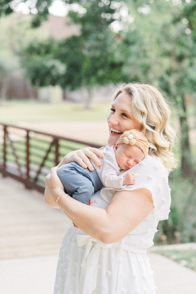 A mother in a white dress holds her newborn baby. The newborn's head is resting on her mother's shoulder. The mother is laughing. Behind them is a bridge and beautiful green trees. This outdoor newborn session is captured by an Austin Newborn Photographer. 