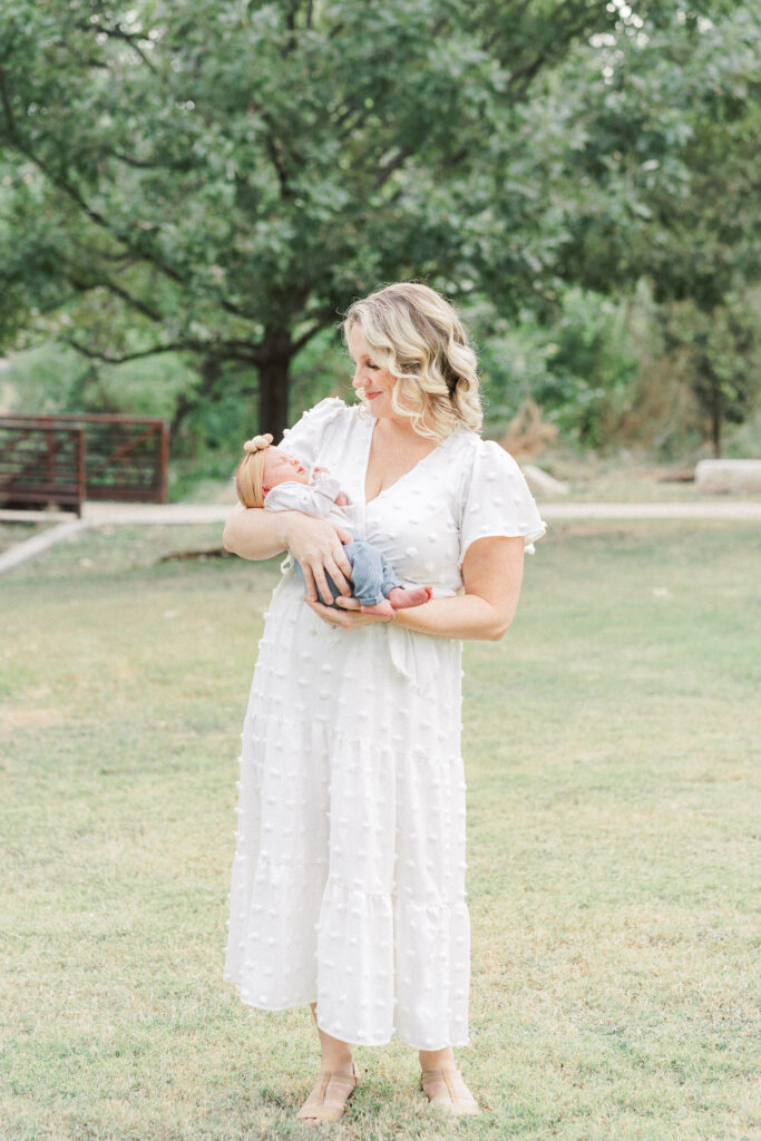 A mother in a long white dress stands in a park while rocking her newborn baby. She looks down at her baby smiling in this authentic moment beautifully captured by an Austin Newborn Photographer.