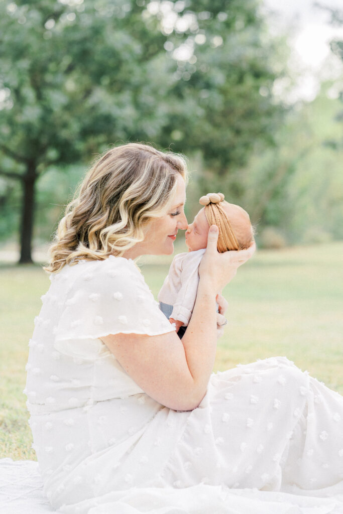 A mother dressed in a long white dress sits on a white blanket in a park while holding her newborn baby. She rubs her nose against her baby's nose while softly smiling in a tender moment captured by an Austin newborn photographer.