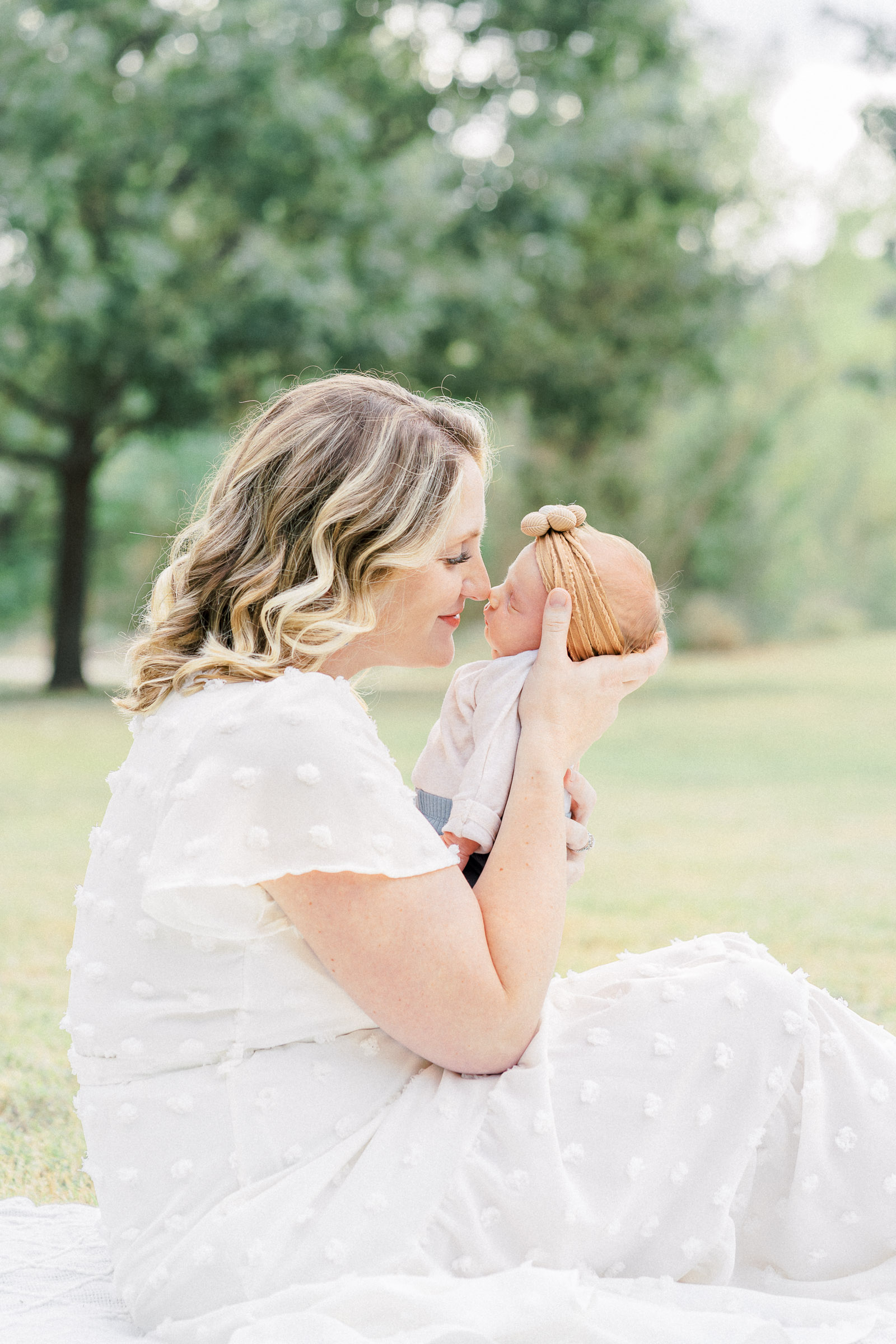 A mother dressed in a long white dress sits on a white blanket in a park while holding her newborn baby. She rubs her nose against her baby's nose while softly smiling in a tender moment captured by an Austin newborn photographer.