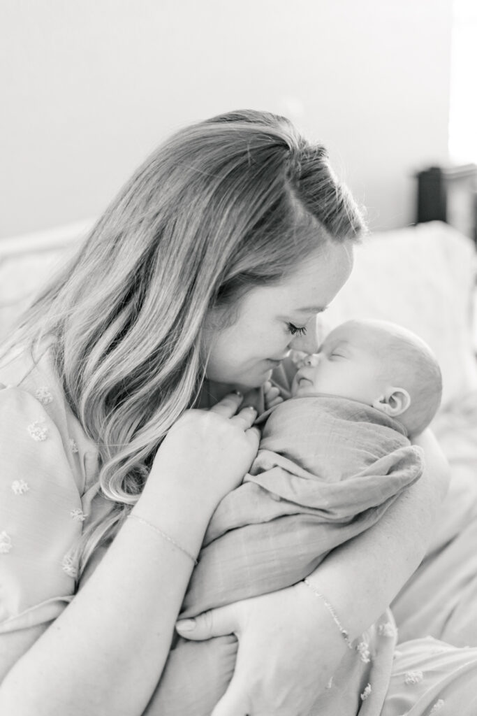 A mother sits on a bed while holding her newborn baby. The baby is swaddled in a blanket. The mother rubs her nose against her baby's nose in this sweet and authentic moment photographed by an Austin newborn photographer.