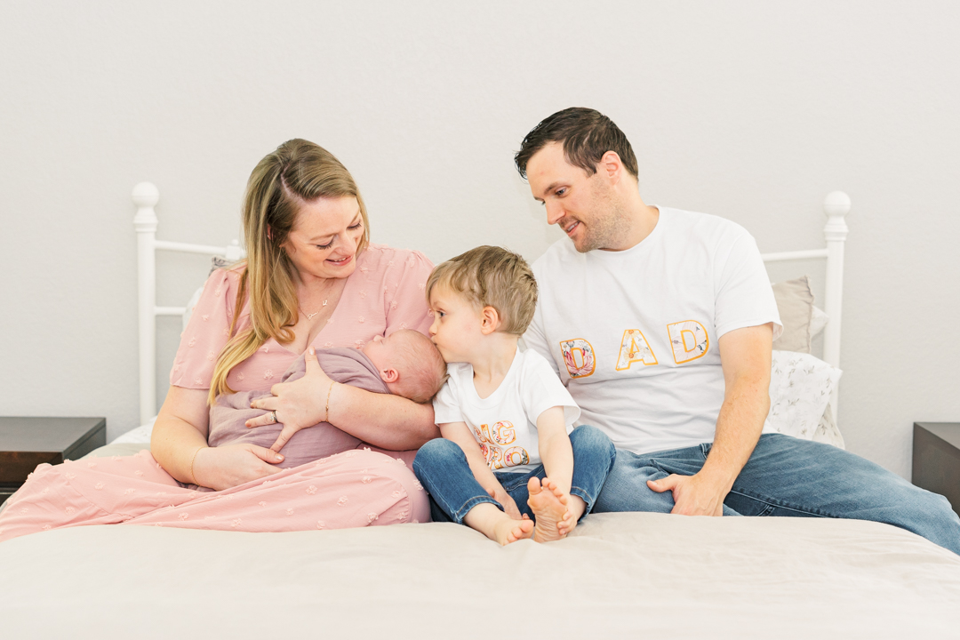 A family sits on a bed, enveloped in a light and airy atmosphere. The mother holds a newborn, while the young boy between the parents gazes naturally at the baby. The father, in his "DAD" shirt, smiles warmly at the authentic scene softly lit around them.