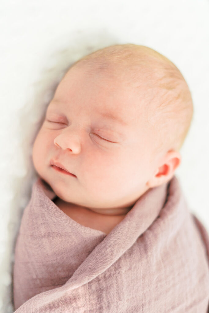 A newborn baby girl is peacefully sleeping, wrapped in a mauve muslin blanket with the sun shining on the left sight of her during a lifestyle newborn session in Round Rock, TX.